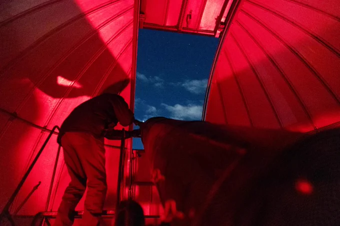 At the top of a ladder in the observatory dome lit by red light, a student looks through the spotting scope of the Stephens Telescope, pointed toward the dark blue night sky through a rectangular opening on Jan. 15, 2025.