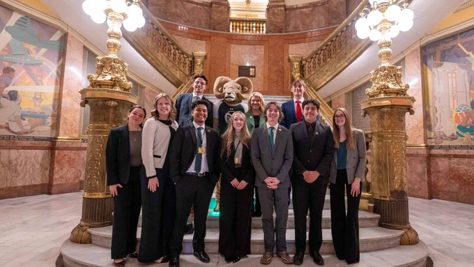 President Amy Parsons stands with A S C S U students and CAM the Ram on the grand staircase inside the Colorado Capitol. The students, dressed in business attire, are smiling, with the historic building’s gold railings, murals, and decorative lamps in the background.