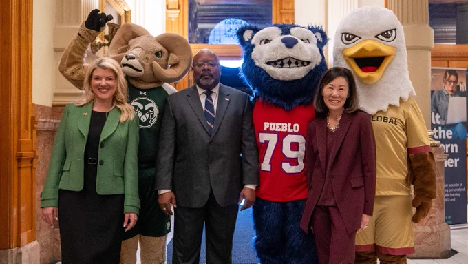 President Amy Parsons stands with university leaders and three mascots—CAM the Ram, the CSU Pueblo ThunderWolf, and the CSU Global eagle—inside the Colorado Capitol. The group is positioned in front of the room’s ornate red walls and gold-trimmed columns, with a CSU representative taking a photo nearby.