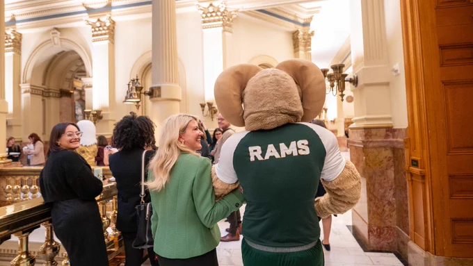 President Amy Parsons walks through the Colorado Capitol with CAM the Ram, smiling. CAM’s jersey reads “RAMS” on the back. Other attendees, including the CSU Global eagle mascot, are visible in the background, along with gold-accented railings and high ceilings.