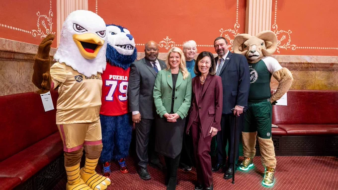 President Amy Parsons stands with university leaders and three mascots—CAM the Ram, the CSU Pueblo ThunderWolf, and the CSU Global eagle—inside the Colorado Capitol. The group is positioned in front of the room’s ornate red walls and gold-trimmed columns, with a CSU representative taking a photo nearby.