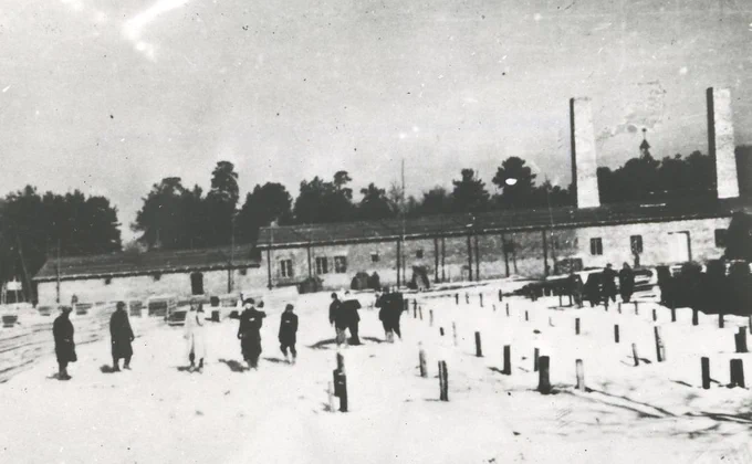 Historical black and white photo of Auschwitz II-Birkenau concentration camp showing prisoners in a snowy field with a building of gas chamber and crematorium IV in the background.
