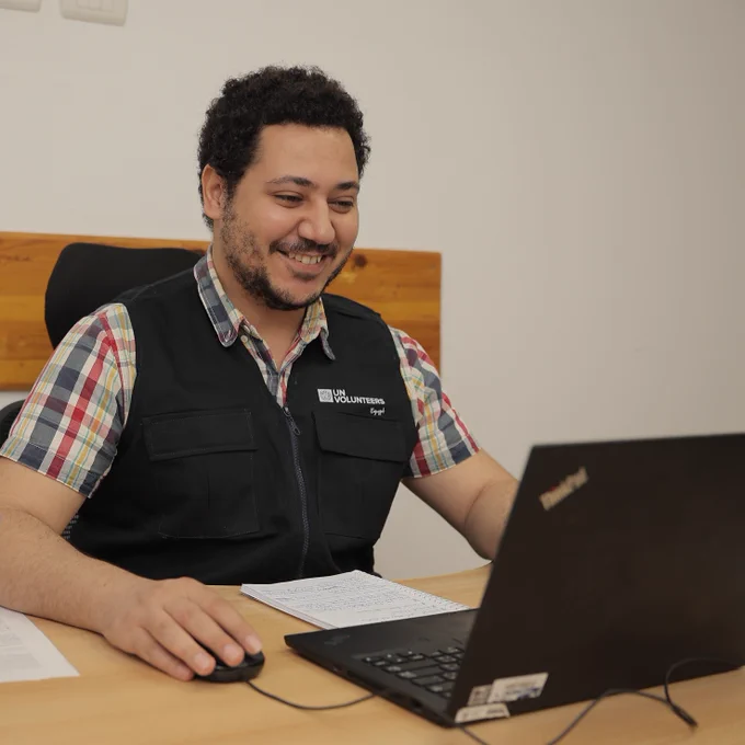  A smiling Volunteer wearing a plaid shirt and a navy blue vest with the “UN Volunteers” logo sits at a desk, working on a laptop. Papers and a pen are on the table, and a blue UN flag is partially visible in the foreground. The setting appears to be an office, and the volunteer looks engaged and focused on his task