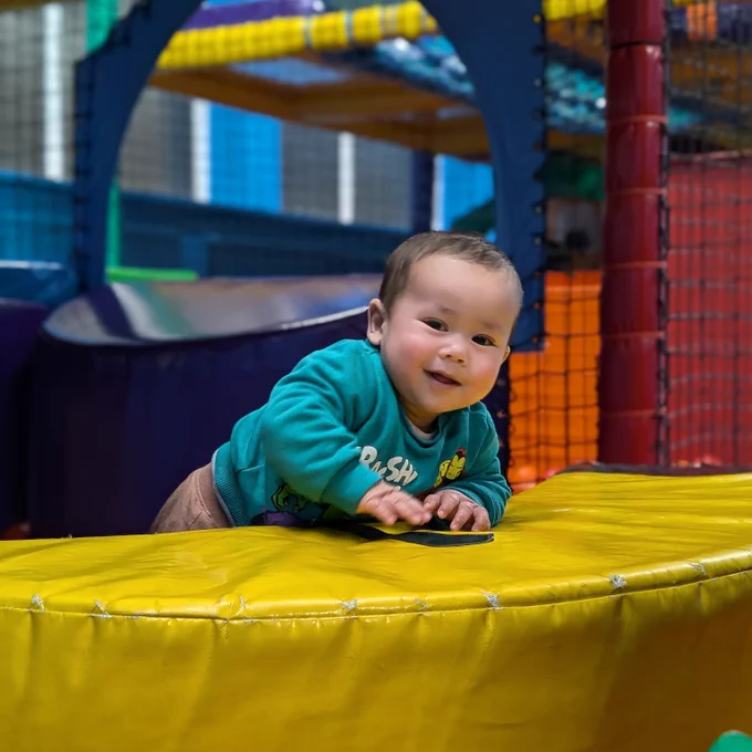 baby ismail in a softplay centre, with a green jumper on, climbing on a yellow cushion and looking to camera 