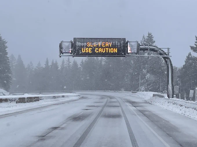 Electronic sign over freeway partially covered in snow says "Slipper, use caution." The road is covered with a thin blank of snow and it is actively snowing.
