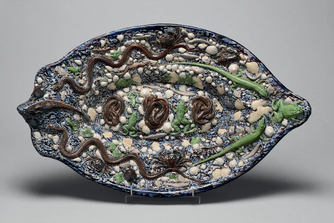 Photo of a piece of ceramic art: an oval dish in dark blue studded with white and cream seashells, decorated with brown ceramic snakes, crabs, and fish, and green ceramic lizards and frogs.