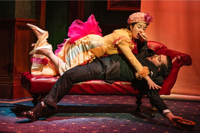 An actress and an actor in period dress wrestle on a chaise lounge on set