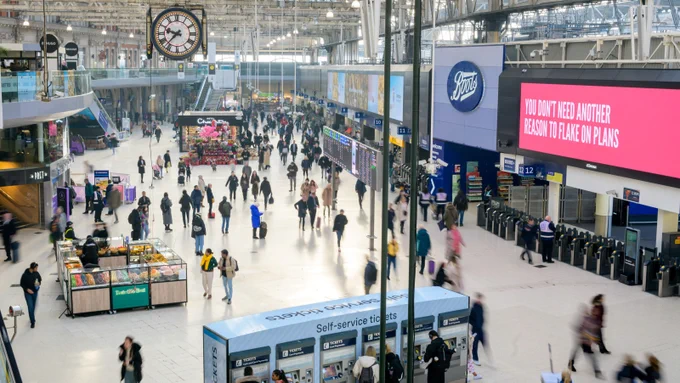 Interior of London Waterloo station with bustling crowd, featuring prominent clocks, ticket machines, and an advertisement for Boots on a large digital screen.