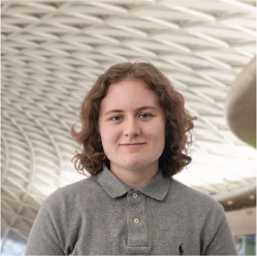 Portrait of Gabrielle, an apprentice in the Property team at Network Rail, wearing a grey polo shirt, standing in front of the intricate roof structure of London King's Cross Station.
