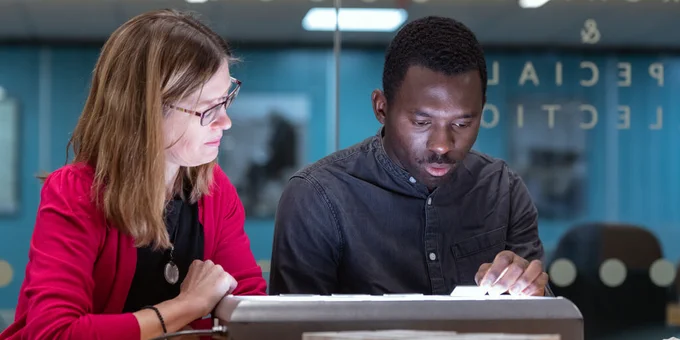 Two students are looking at an archival lightbox. One is wearing glasses and the other is wearing a black shirt.