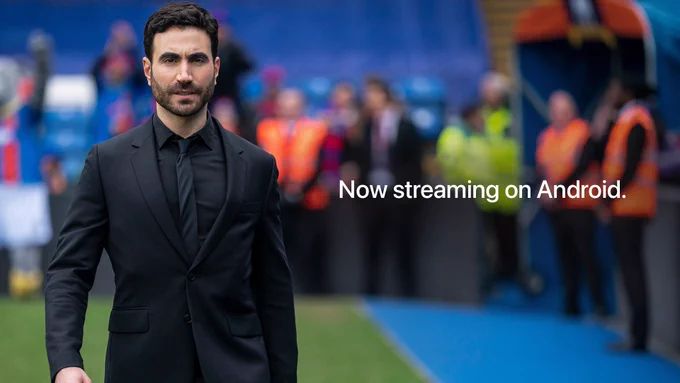 From Ted Lasso. Roy, portrayed by Brett Goldstein, serves in an all-black suit and tie. He is standing on a field with various spectators behind him. There is text on-screen that reads "Now streaming on Android."