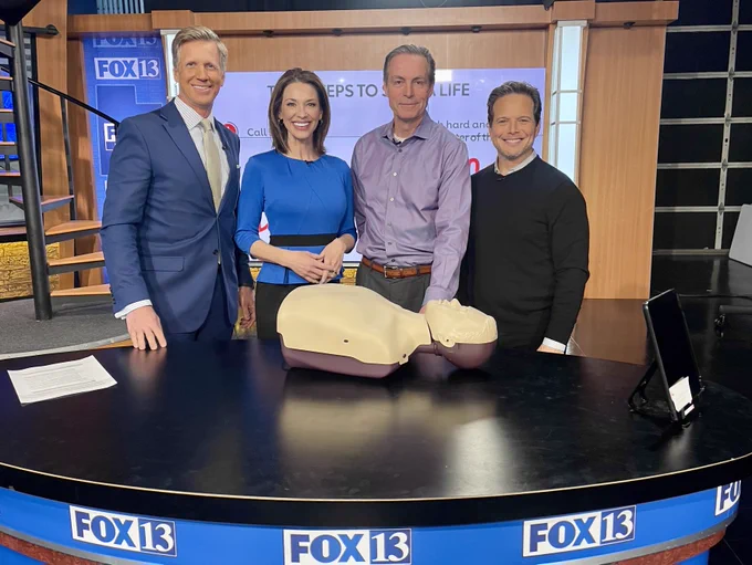 FOX13 Good Day Utah anchors, Dan Evans and Kelly Chapman, stand next to MountainStar Healthcare Chief Medical Officer, Dr. Filip Roos, on set, smiling. On the other side of Dr. Roos is actor, Scott Wolf, who plays a physician in a new medical drama on FOX entitled, “Doc.” A CPR manikin lays flat on the table in front of the group.