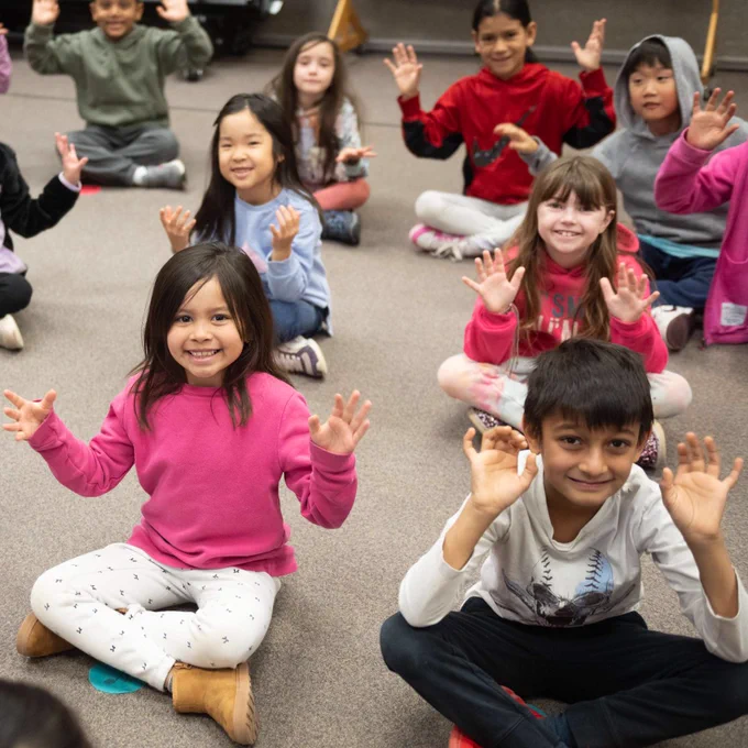 Elementary students sit on a carpet in a classroom, holding their hands up and open while smiling.