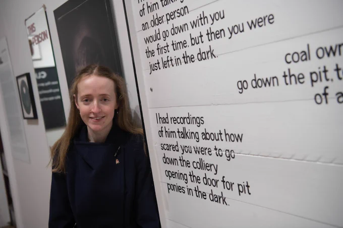 A person with long dark blonde hair and a dark navy coat standing in front of a white and black painted sign.