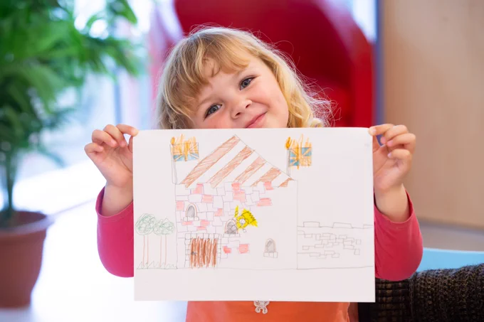 A child with blonde curly hair and a pink and orange T-shirt. They are smiling and holding up a colourful drawing on a white piece of paper.