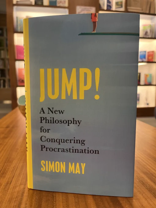 A hardback copy of Jump! by Simon May, propped on a table with a display of bookshelves in the background. The cover shows the bottom half of a person preparing to jump from a diving board, against the backdrop of a blue sky with bold yellow text. 