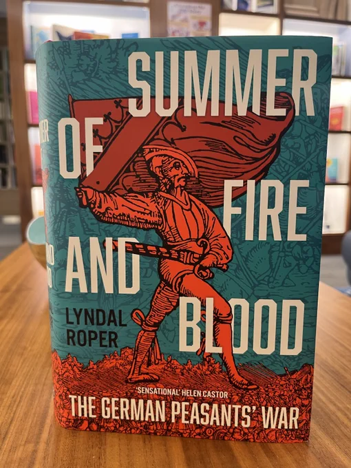 A hardback copy of Summer of Fire and Blood by Lyndal Roper, propped on a table with a display of bookshelves in the background. The cover shows an illustration of a bright red figure carrying a flag and wielding a sword, against a backdrop of an illustrated battle scene.