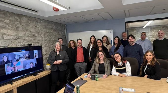 Team members smiling from Compass Rose in a "family photo" at our office in our boardroom. There is a screen which also has people smiling who are on Zoom. 