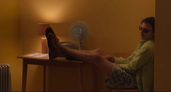 Person wearing sunglasses and shorts sitting with feet on a table, next to a fan and a radiator in a room with warm lighting.