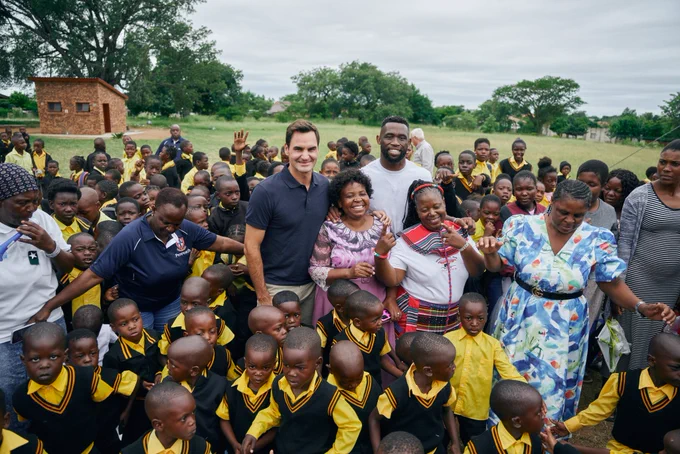 Roger Federer and Siya Kolisi surrounded by uniformed preschool children and staff. A green field and trees can be seen in the background.