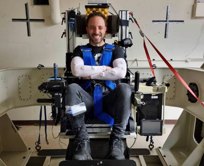 Dr Aaron Parkhurst (UCL Anthropology) received a Social Science Plus award to support the Off-World Living project. In the photo, he is smiling while seated in a specialized chair with blue harnesses and arm restraints, surrounded by equipment in a room with wall-mounted devices. The project explores analogue environments designed for off-world living. 