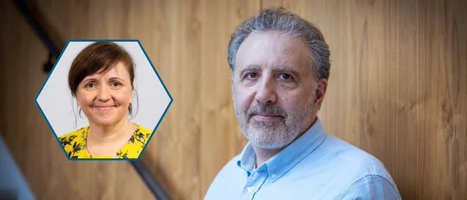 A man with short grey hair and a beard stands against a wooden panel background, wearing a light blue shirt. An inset hexagonal frame in the foreground contains a portrait of a woman with short brown hair, wearing a yellow floral blouse and smiling
