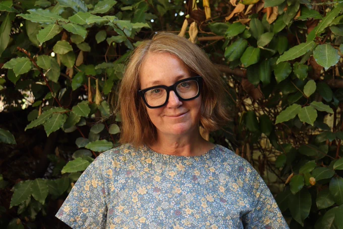 Photograph of author Judith Eagle. She is in front of green foliage, looking at the camera and smiling, she wears a floral top and very stylish black-framed glasses.