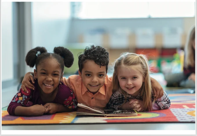 Three happy, children lying down on a brightly coloured mat with an open book, smiling into the camera.