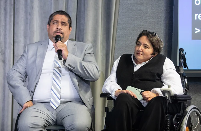 The image shows Jose Maria Viera and Ola Abualghaib sitting side by side.
Mr Viera in a light grey suit with a striped tie speaks through a microphone.
Ms. Abualghaib wearing a white shirt and black vest listens attentively.
The background is a neutral setting with grey curtains and a screen displaying text.