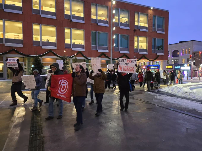 A picture of protestors walking to city hall in Kitchener. The red clay panels in the back ground of a couple dozen people marching with signs. Photo: WR ACORN