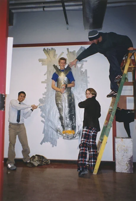 A man is fully taped to a white wall with duct tape, while three students stand around him and point at him. One student is standing on a ladder and pointing down at him.