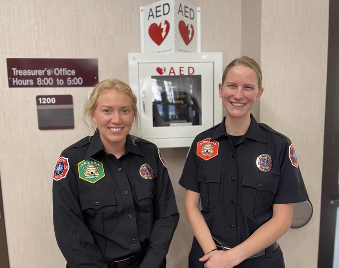 Two members of the Spanish Fork Fire Department’s EMS team pose together, smiling, beside an AED posted up at the Utah County Government public building.