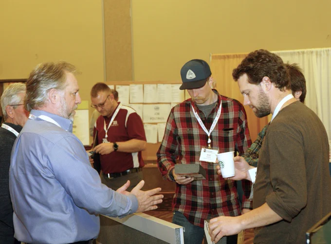 Group of individuals engaging in a discussion at a conference booth, with some holding brochures and digital devices.