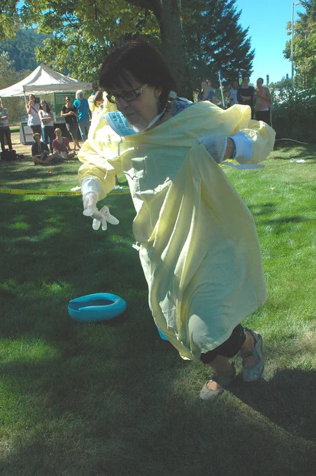 A woman wearing scrubs and gloves runs outside on the grass. A blue bed pan is seen in the background along with a group of people.