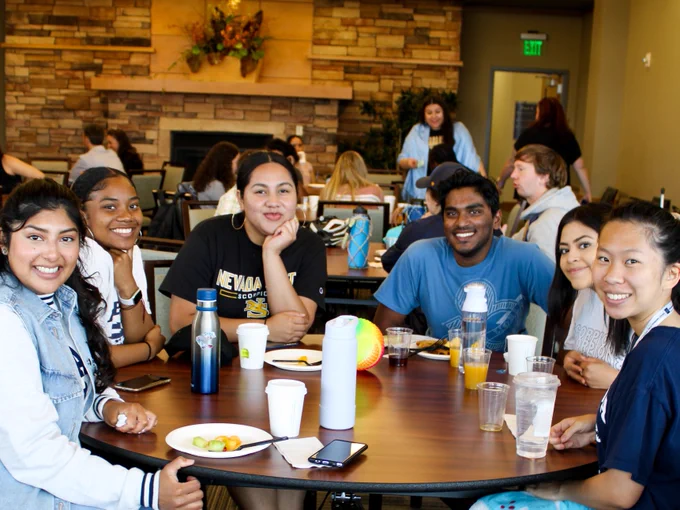 A group of students sitting around a table, enjoying a meal together at the University of Nevada. Some beverages and food items are on the table. They are smiling and engaged in conversation in a room with a stone fireplace in the background.