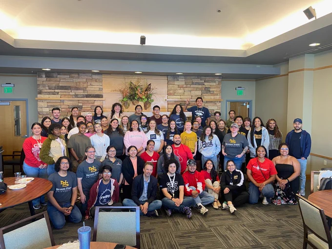 Group of people smiling in a large room, gathered for an event, possibly a community or university gathering, with a natural light setting and modern decor in the background.