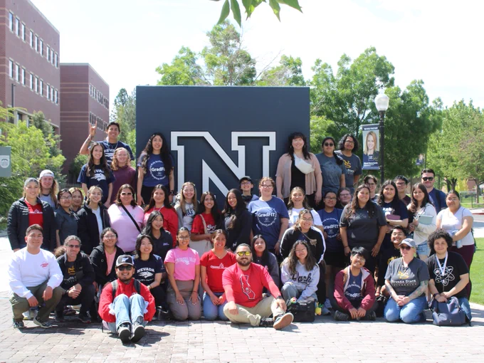 A group of people posing in front of a large block letter N, likely representing a university or college, on a sunny day with trees and buildings in the background.
