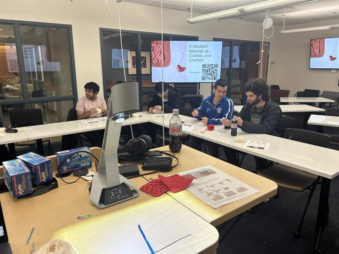 A group of students learning crochet. A slide reads “Cookies and Crochet for VAL-entine’s Day”