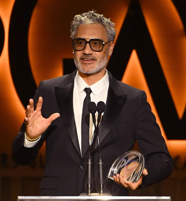 Taika stands in a black and white suit at the Producers Guild with a orange background