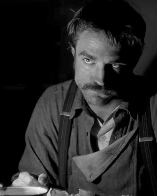 Thomas Howard (Robert Pattinson) looking intently with a moustache and shirt and dungarees 