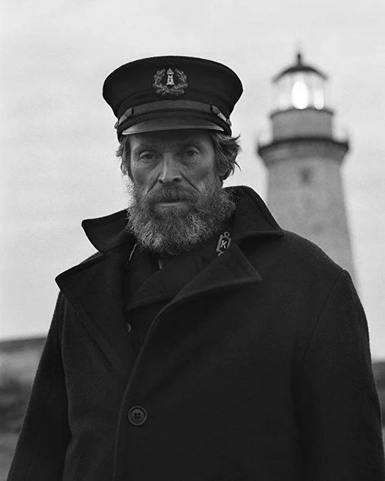 Thomas Wake (Willem Defoe) with a big bushy beard, sailors hat and big coat with the lighthouse in the background 