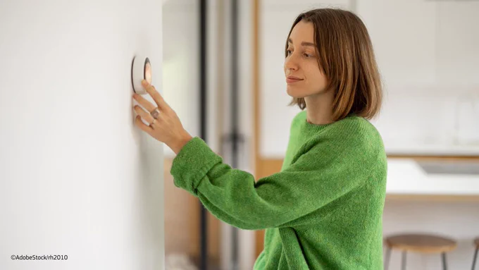 Young woman wearing a green jumper turning down the thermostat in the living room. 