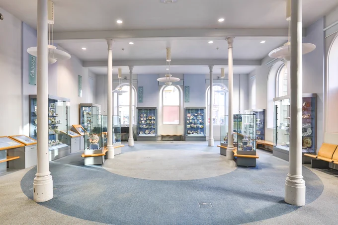 A pale lilac and blue gallery space with a blue and lilac carpet in circular bands. The room has six pillars from floor to ceiling. It also has glass display cases with a selection of different shaped pottery.