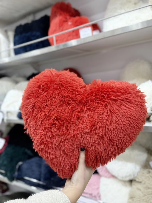 A person's hand holding a large, plush red heart-shaped pillow in a store aisle filled with various cushions and soft toys.