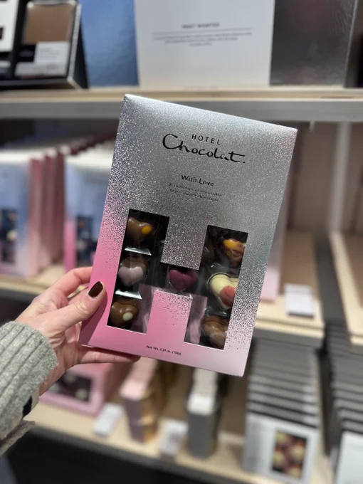 A hand holding a box of Hotel Chocolat assorted chocolates with a 'With Love' inscription, displayed in a store.