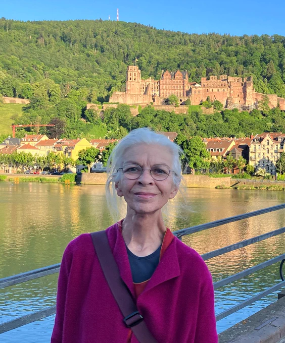 Gabriele smiles outdoors in Heidelberg. There is a body of water behind her and several buildings beyond the water nestled on a landscape amongst many green trees.