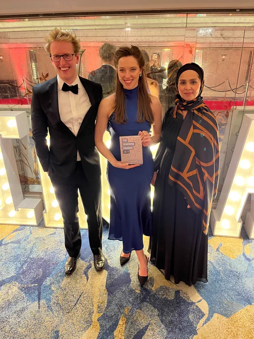 Three individuals pose for a photo at an event, with one holding an award. The person on the left is dressed in a black tuxedo, the middle person in a blue gown, and the one on the right in a black and orange hijab and dress. They are standing in a brightly lit venue with a reflective floor.