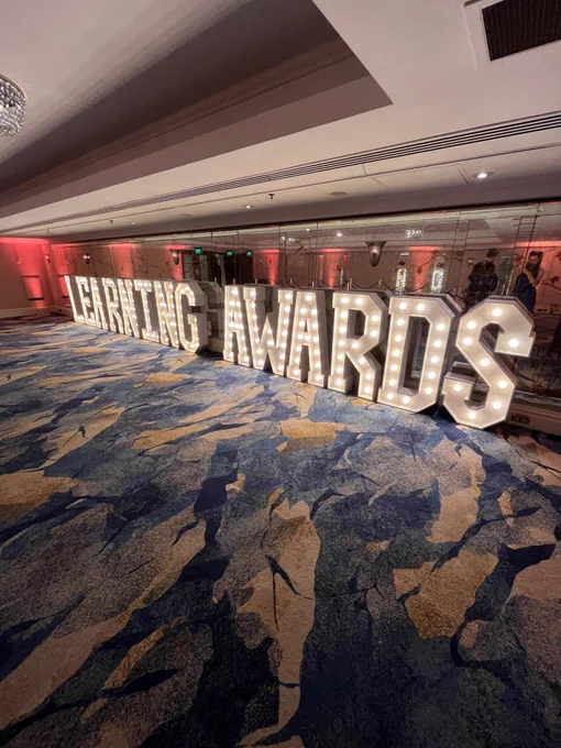 Large illuminated letters spelling "LEARNING AWARDS" on the floor of a carpeted hallway with decorative lighting.