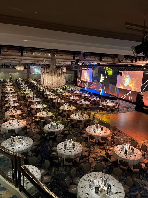 Elegant event setup in a grand hall featuring round dining tables, each set with white tablecloths and wine glasses, before a large stage with vibrant, illuminated screens displaying abstract designs. A large chandelier hangs overhead.