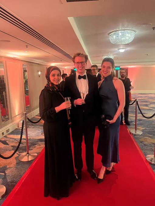 Three people dressed in formal attire standing on a red carpet at an event.
